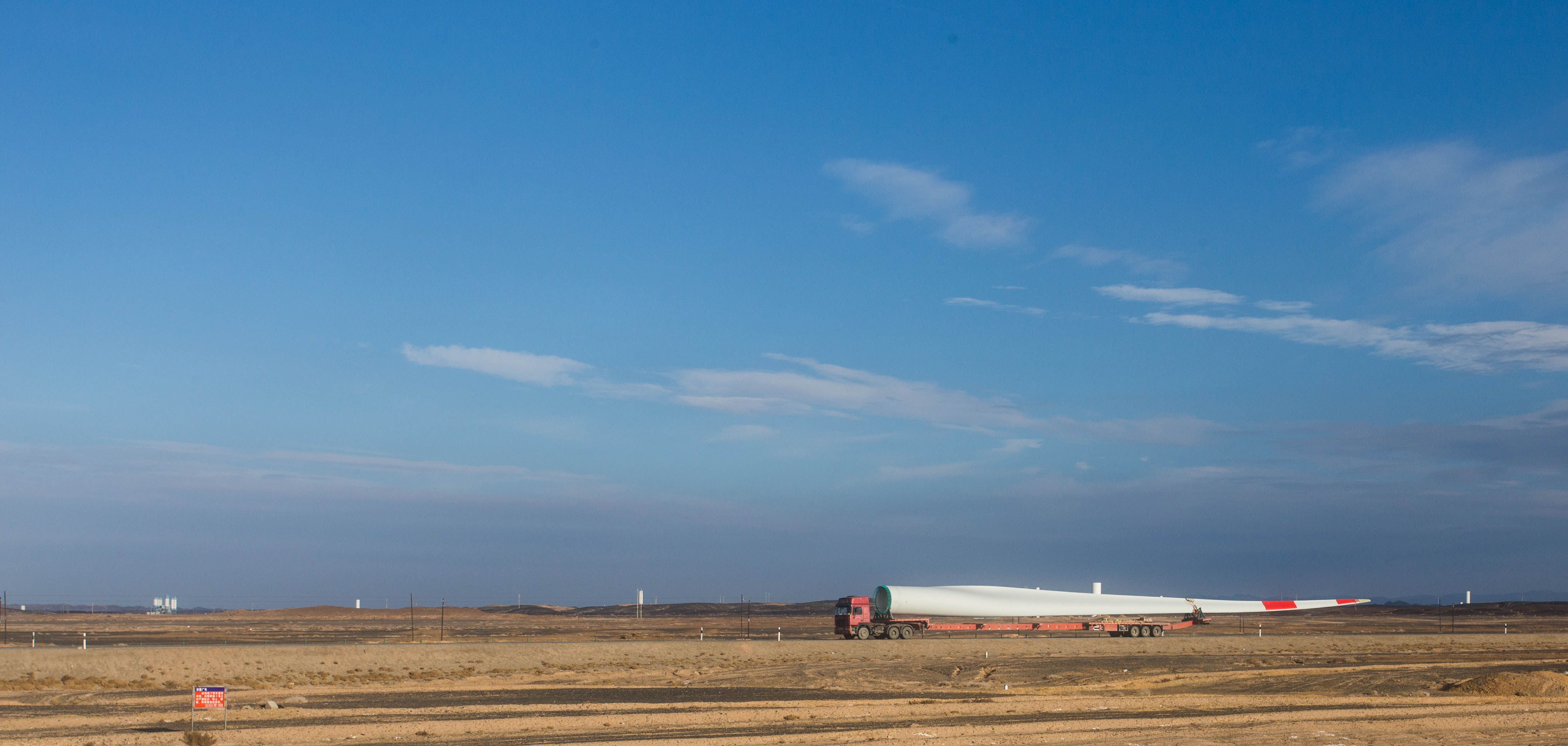 The blade of a wind turbine being transported across the vast desert plains of western China
